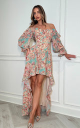 A woman models a floral off-the-shoulder high-low dress with long bell sleeves, ruffled trim, and a sheer lining, standing against a white paneled wall.